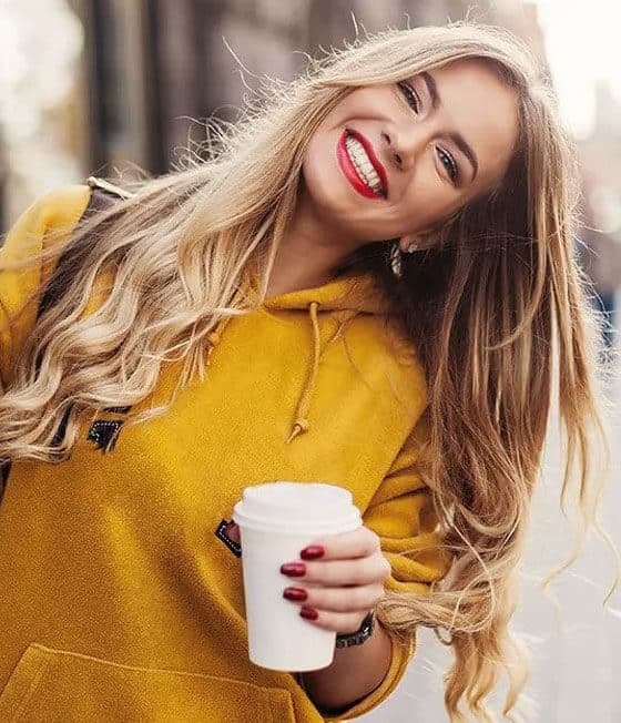 Smiling woman in yellow jacket holding coffee cup, displaying bright white teeth and red lipstick.